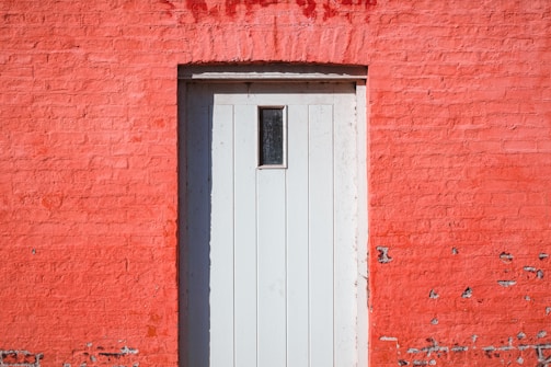 A wooden door with vertical panels set into a vivid red brick wall. The door is painted white and features a small rectangular window at the top. There are some signs of wear and tear on the wall, with spots where the paint has chipped away.