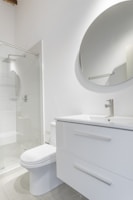 A modern bathroom interior featuring a white vanity with a sleek basin and a silver faucet. Above the vanity is a large round mirror. To the left, there's a glass-enclosed shower with a silver showerhead. The walls and floor have a minimalistic design with light, neutral colors.