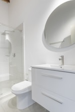 A modern bathroom interior featuring a white vanity with a sleek basin and a silver faucet. Above the vanity is a large round mirror. To the left, there's a glass-enclosed shower with a silver showerhead. The walls and floor have a minimalistic design with light, neutral colors.