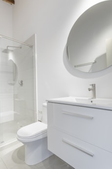 A modern bathroom interior featuring a white vanity with a sleek basin and a silver faucet. Above the vanity is a large round mirror. To the left, there's a glass-enclosed shower with a silver showerhead. The walls and floor have a minimalistic design with light, neutral colors.