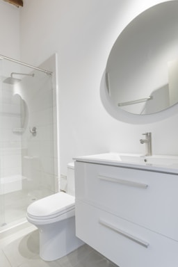 A modern bathroom interior featuring a white vanity with a sleek basin and a silver faucet. Above the vanity is a large round mirror. To the left, there's a glass-enclosed shower with a silver showerhead. The walls and floor have a minimalistic design with light, neutral colors.
