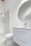 A modern bathroom interior featuring a white vanity with a sleek basin and a silver faucet. Above the vanity is a large round mirror. To the left, there's a glass-enclosed shower with a silver showerhead. The walls and floor have a minimalistic design with light, neutral colors.