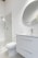 A modern bathroom interior featuring a white vanity with a sleek basin and a silver faucet. Above the vanity is a large round mirror. To the left, there's a glass-enclosed shower with a silver showerhead. The walls and floor have a minimalistic design with light, neutral colors.