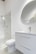 A modern bathroom interior featuring a white vanity with a sleek basin and a silver faucet. Above the vanity is a large round mirror. To the left, there's a glass-enclosed shower with a silver showerhead. The walls and floor have a minimalistic design with light, neutral colors.