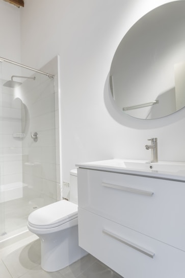 A modern bathroom interior featuring a white vanity with a sleek basin and a silver faucet. Above the vanity is a large round mirror. To the left, there's a glass-enclosed shower with a silver showerhead. The walls and floor have a minimalistic design with light, neutral colors.