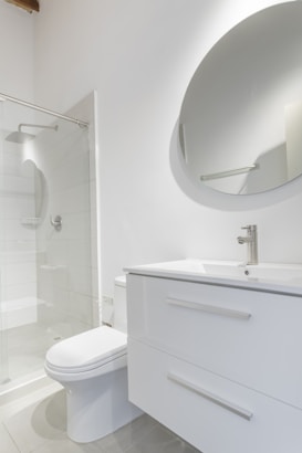 A modern bathroom interior featuring a white vanity with a sleek basin and a silver faucet. Above the vanity is a large round mirror. To the left, there's a glass-enclosed shower with a silver showerhead. The walls and floor have a minimalistic design with light, neutral colors.