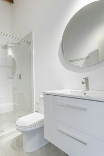 A modern bathroom interior featuring a white vanity with a sleek basin and a silver faucet. Above the vanity is a large round mirror. To the left, there's a glass-enclosed shower with a silver showerhead. The walls and floor have a minimalistic design with light, neutral colors.