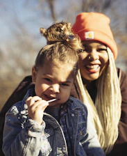 A young child with curly hair wearing a denim jacket holds a lollipop. In the background, an adult with long light-colored braids and an orange beanie smiles warmly.
