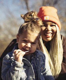 A young child with curly hair wearing a denim jacket holds a lollipop. In the background, an adult with long light-colored braids and an orange beanie smiles warmly.