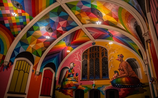 A vibrantly painted interior featuring a high ceiling adorned with geometric patterns in a spectrum of colors. The walls display large architectural windows, and colorful murals depicting abstract figures sitting beneath trees. Artwork combines both modern and traditional elements with a focus on symmetry and rich detail.