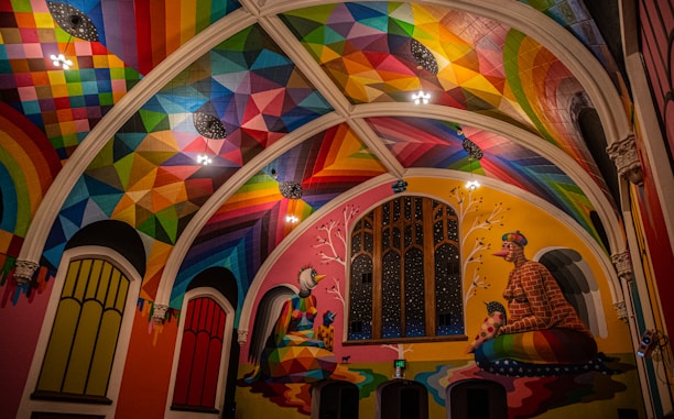 A vibrantly painted interior featuring a high ceiling adorned with geometric patterns in a spectrum of colors. The walls display large architectural windows, and colorful murals depicting abstract figures sitting beneath trees. Artwork combines both modern and traditional elements with a focus on symmetry and rich detail.