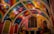 A vibrantly painted interior featuring a high ceiling adorned with geometric patterns in a spectrum of colors. The walls display large architectural windows, and colorful murals depicting abstract figures sitting beneath trees. Artwork combines both modern and traditional elements with a focus on symmetry and rich detail.