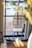 A cozy, bright room features a wicker-style chair with white cushions positioned in front of large windows. The windows offer a view of the outdoors with bare trees visible, suggesting a seasonal change. A soft rug lies on the floor beneath the chair, adding a homely touch.