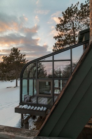 A modern house with large glass windows surrounded by a snowy landscape and pine trees under a sky filled with soft clouds. The architectural design features sharp angles and a small overhang with snow accumulation.