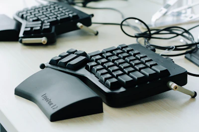 A custom ergonomic keyboard with a unique split design rests on a desk. The keyboard features mechanical keys with an extended wrist support, surrounded by connecting cables.