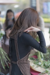 A person is engaged in flower arranging, surrounded by various stems and leaves. They are wearing a dark ribbed top and a brown apron. The background is slightly blurred, suggesting a busy or outdoor setting with other people around.