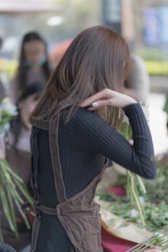 A person is engaged in flower arranging, surrounded by various stems and leaves. They are wearing a dark ribbed top and a brown apron. The background is slightly blurred, suggesting a busy or outdoor setting with other people around.