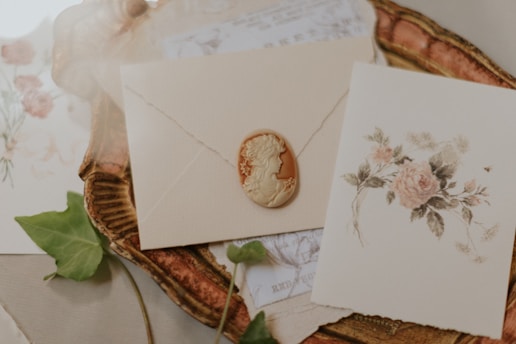 A vintage-style arrangement featuring an embossed envelope with a cameo seal, a card with a floral illustration, and a decorative tray. The scene includes delicate paper details and a green leaf on the side.