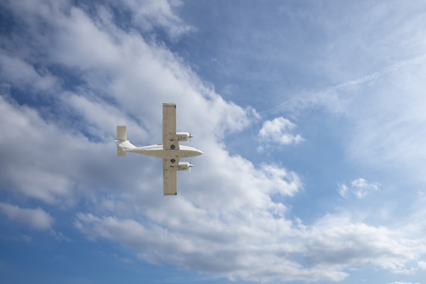 A light aircraft is flying through a partly cloudy blue sky. The plane is central in the image with its wingspan visible, set against a backdrop of scattered white clouds.