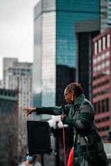 A person with braided hair and sunglasses is speaking into a microphone, gesturing with a pointed hand. They are wearing a checkered jacket and standing in front of a backdrop of tall buildings. A speaker is visible beside them, and another individual wearing a mask stands nearby.