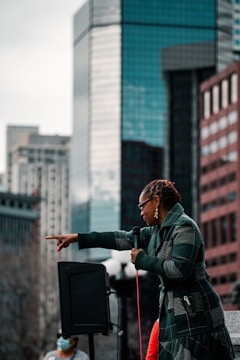 A person with braided hair and sunglasses is speaking into a microphone, gesturing with a pointed hand. They are wearing a checkered jacket and standing in front of a backdrop of tall buildings. A speaker is visible beside them, and another individual wearing a mask stands nearby.