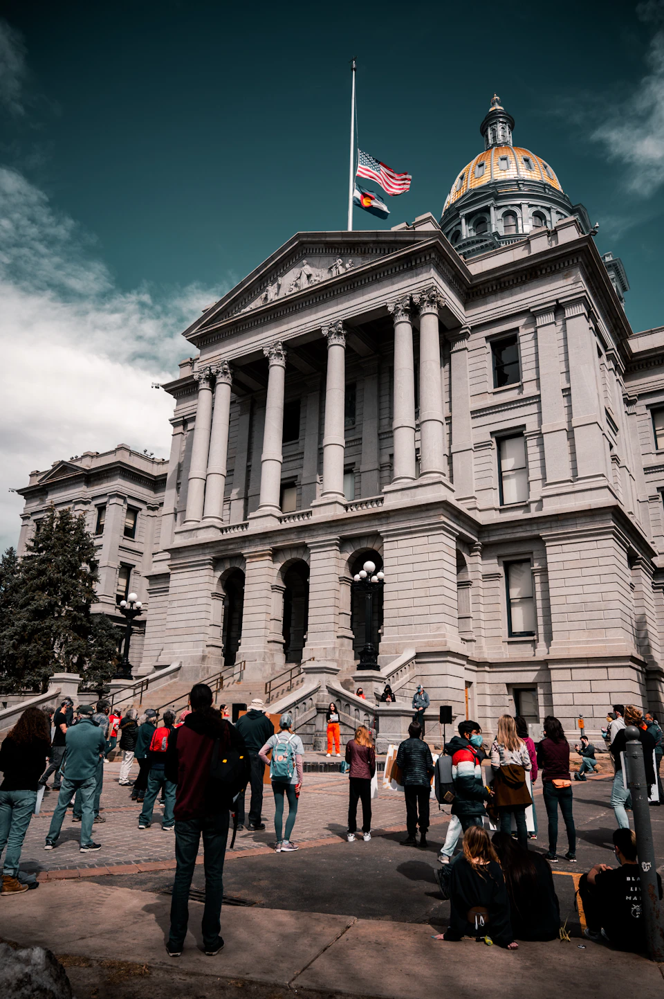 The image depicts a large, white stone building with columns and a dome, likely a courthouse or government building. A flagpole stands tall on top of the structure, fluttering in the breeze. The building is surrounded by trees, providing a natural backdrop to its architectural grandeur. In front of the building, there are several people gathered, some standing and others sitting, possibly waiting for an event or simply enjoying their surroundings.