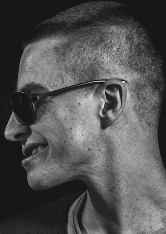 A portrait captures a person in profile, turned to the side, wearing sunglasses. The lighting emphasizes the texture of the skin and creates a contrast with the dark background.