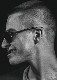 A portrait captures a person in profile, turned to the side, wearing sunglasses. The lighting emphasizes the texture of the skin and creates a contrast with the dark background.