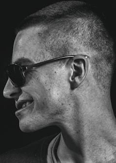 A portrait captures a person in profile, turned to the side, wearing sunglasses. The lighting emphasizes the texture of the skin and creates a contrast with the dark background.