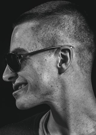 A portrait captures a person in profile, turned to the side, wearing sunglasses. The lighting emphasizes the texture of the skin and creates a contrast with the dark background.