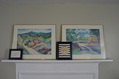 Two framed landscape paintings are displayed on a white mantel. The paintings are colorful, depicting serene natural scenes with trees and paths. Two smaller framed prints are placed in front of the paintings. One features the word 'Love' in cursive, and the other has the text 'Happy Place' against a striped background.