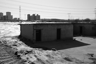 A barren landscape with two small, dilapidated buildings in the foreground, surrounded by a deserted area. In the background, there are high-rise buildings and electricity pylons, suggesting a contrast between urban development and rural decay.