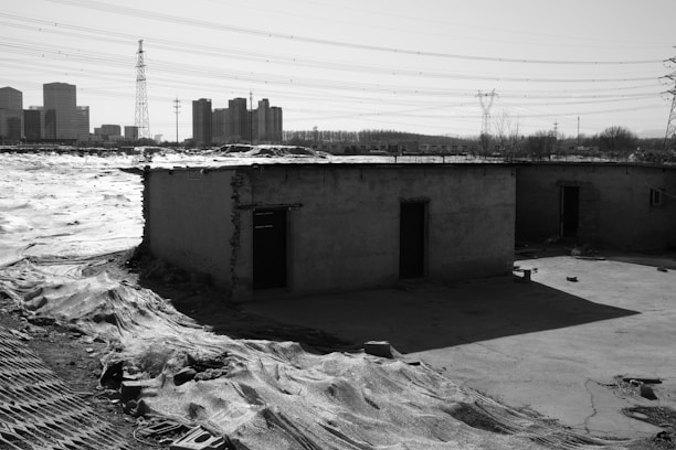A barren landscape with two small, dilapidated buildings in the foreground, surrounded by a deserted area. In the background, there are high-rise buildings and electricity pylons, suggesting a contrast between urban development and rural decay.