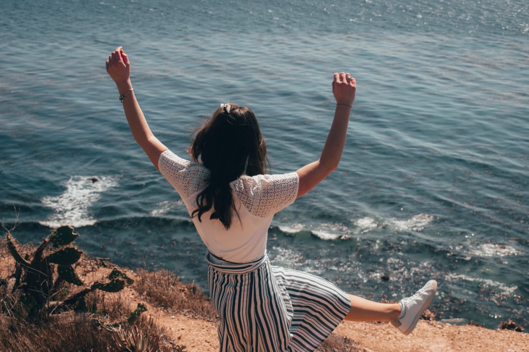 woman in white and black stripe dress standing on brown rock near body of water during