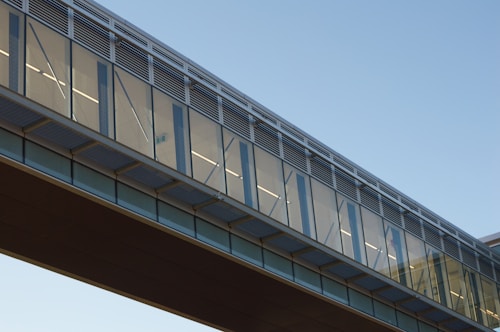 A modern skybridge with a sleek design features large glass panels and metallic frames. The structure is bathed in daylight under a clear blue sky, and reflections on the glass add a dynamic visual element.