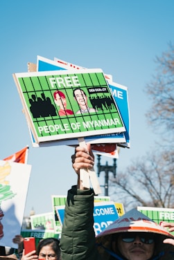 A protestor holds several signs demanding freedom for the people of Myanmar. The signs are bright green with illustrations and messages. Other individuals are partially visible, one wearing a traditional hat.