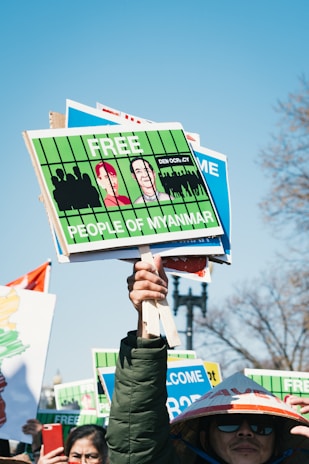A protestor holds several signs demanding freedom for the people of Myanmar. The signs are bright green with illustrations and messages. Other individuals are partially visible, one wearing a traditional hat.