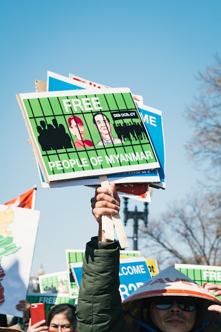 A protestor holds several signs demanding freedom for the people of Myanmar. The signs are bright green with illustrations and messages. Other individuals are partially visible, one wearing a traditional hat.