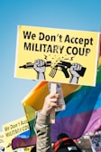 A person holds a vibrant protest sign that reads 'We Don't Accept Military Coup' with a graphic of two hands breaking a rifle, set against a clear blue sky. The background features colorful rainbow flags and other signs, indicating a protest or demonstration setting.