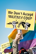 A person holds a vibrant protest sign that reads 'We Don't Accept Military Coup' with a graphic of two hands breaking a rifle, set against a clear blue sky. The background features colorful rainbow flags and other signs, indicating a protest or demonstration setting.