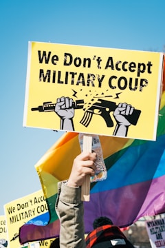 A person holds a vibrant protest sign that reads 'We Don't Accept Military Coup' with a graphic of two hands breaking a rifle, set against a clear blue sky. The background features colorful rainbow flags and other signs, indicating a protest or demonstration setting.