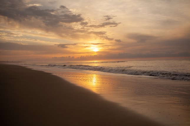 A serene beach at sunset with gentle waves lapping at the shore. The sun is partially obscured by scattered clouds, casting a warm, golden glow across the sky and reflecting on the wet sand.