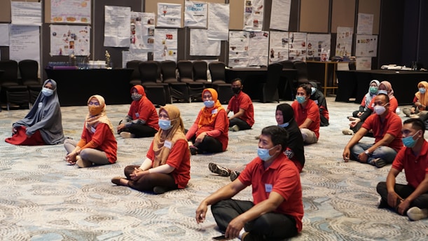 A group of people sits on the floor of a meeting room, wearing face masks and red shirts. The room is spacious with patterned carpeting, and the walls are adorned with displayed charts and posters. Chairs are lined up at the back of the room, and the participants appear attentive and engaged.