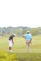 Two individuals are on a golf course. One person is preparing to putt while the other watches with a golf club in hand. They are dressed in typical golf attire, including caps and polo shirts. The landscape is expansive with well-manicured grass, and trees can be seen in the background.