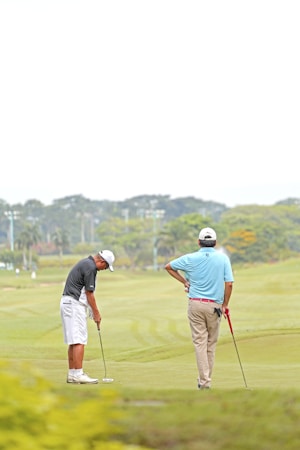 Two individuals are on a golf course. One person is preparing to putt while the other watches with a golf club in hand. They are dressed in typical golf attire, including caps and polo shirts. The landscape is expansive with well-manicured grass, and trees can be seen in the background.