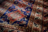 An intricately patterned rug with a dominant blue and red color scheme. The central section displays symmetrical floral designs with geometric shapes interspersed, while the border features a series of repeating motifs in complementary colors.