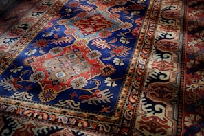 An intricately patterned rug with a dominant blue and red color scheme. The central section displays symmetrical floral designs with geometric shapes interspersed, while the border features a series of repeating motifs in complementary colors.