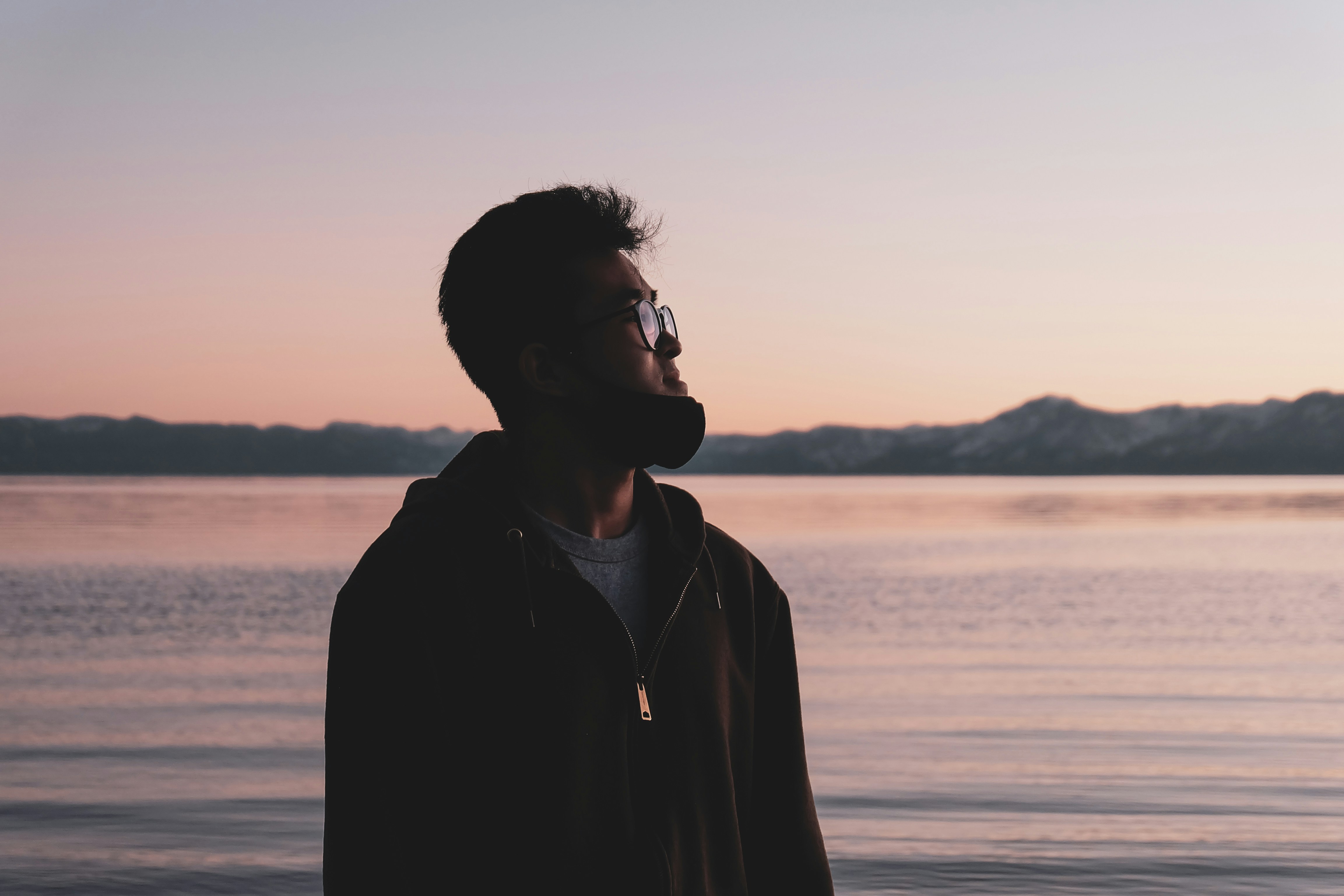 man in black jacket standing near body of water during sunset