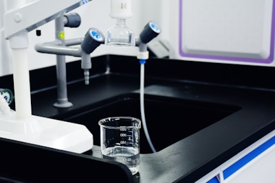 A laboratory workspace features a black countertop with a beaker partly filled with liquid. Above the beaker, there is laboratory equipment including faucets and dispensers. The environment appears sterile and clean, typical of a scientific or research setting.