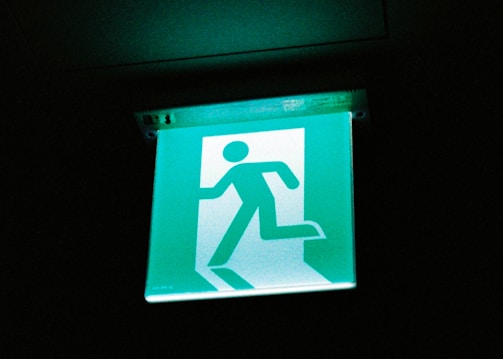 An illuminated emergency exit sign featuring a simplified figure running through a door. The sign is bright green and contrasts sharply against the dark background.